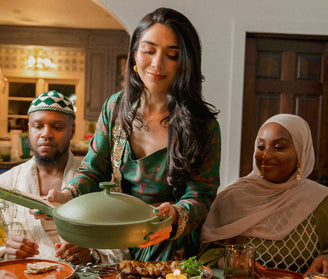 A woman in a green dress serves food from a large green pan at a dinner table, while two guests dressed in traditional attire smile and watch her. The setting appears warm and festive.