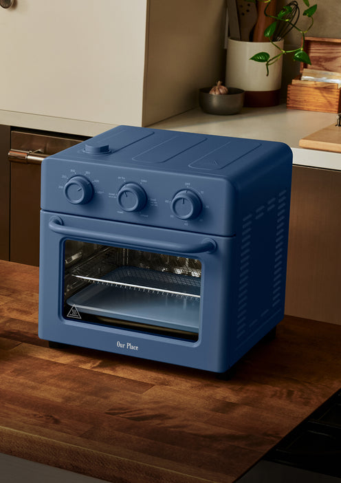 A blue countertop oven with three control knobs is placed on a wooden kitchen counter near a plant and kitchen utensils.