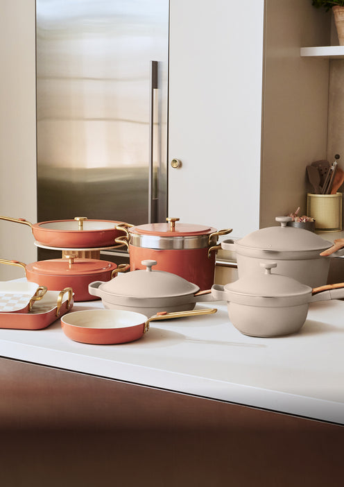Various pots, pans, and lids with gold handles in beige and terracotta colors are arranged on a modern white kitchen counter.