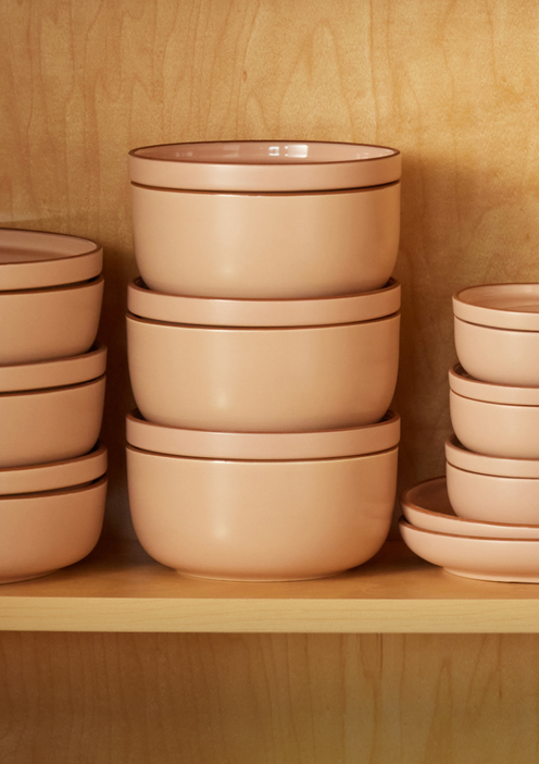 A set of stacked, light pink ceramic bowls and plates are neatly arranged on a wooden shelf with a light wood grain background.