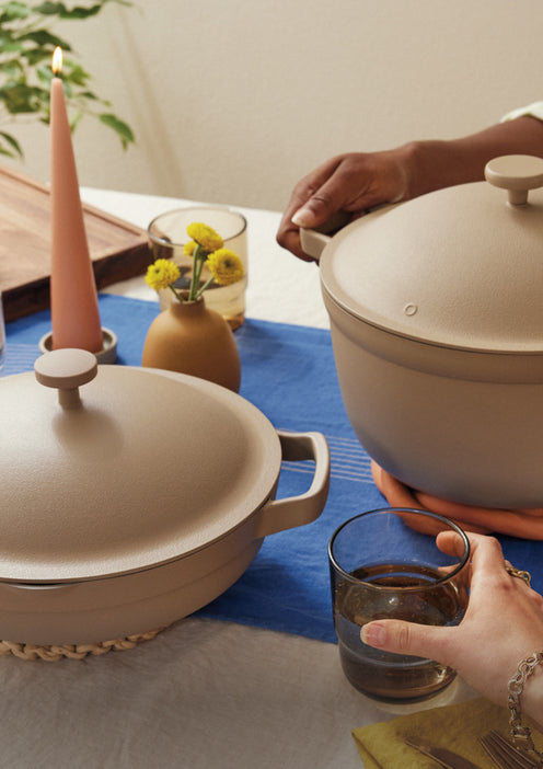 Two hands are shown at a dining table, one lifting the lid off a beige pot, the other holding a glass of water. On the table are another covered pot, a pink candle, yellow flowers in a vase, and a blue table runner.