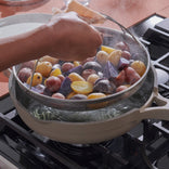 A hand lifts a steamer basket filled with assorted small potatoes, including purple, red, and yellow ones, out of a pot of boiling water on a stovetop. Steam rises from the pot.