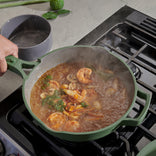 A person cooks a seafood stew with shrimp, mushrooms, and herbs in a green pot on a stovetop. Steam rises from the simmering dish, and a gray bowl is held nearby, ready to serve.