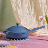 A blue skillet with a matching lid and light wooden handle sits on a round trivet atop a pink surface, with a light pink flower nearby and soft, draped fabric in the background.
