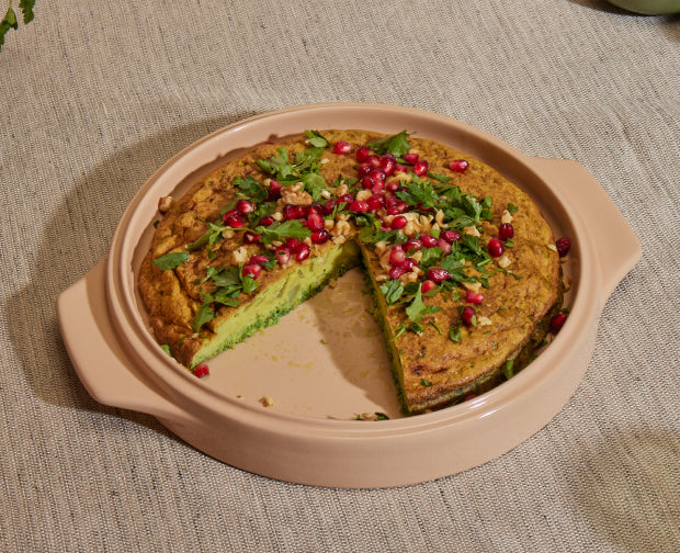 A round herb frittata with a slice missing, topped with chopped herbs, walnuts, and pomegranate seeds, displayed in a beige baking dish on a textured beige tablecloth.