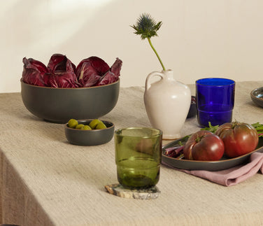 A table set with bowls of radicchio, tomatoes, olives, pickles, seeds, a wedge of cheese, dates, and ceramic pitchers, with green glasses and purple napkins, all arranged in soft natural light.