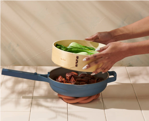 A person places a bamboo steamer with bok choy over a blue pan filled with cooked meat, set on a tiled surface with soft natural lighting.