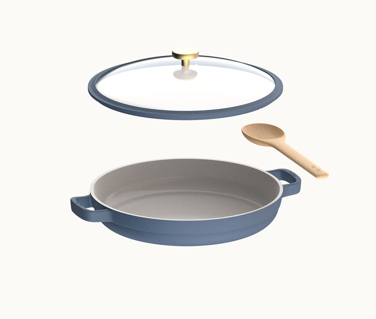 Blue ceramic-coated pan with two handles, accompanied by a clear glass lid with a gold knob and a wooden spoon, all shown on a plain white background.