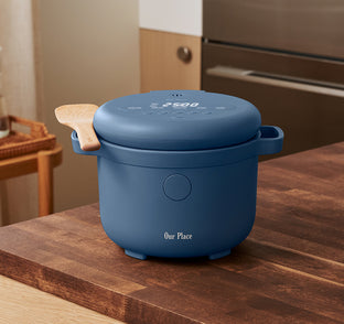 A blue electric rice cooker with a digital display and wooden-handled spoon, labeled Our Place, sits on a wooden kitchen counter.