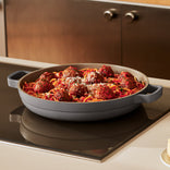 A large gray pan holds spaghetti topped with tomato sauce, meatballs, and grated cheese, set on a stovetop in a modern kitchen.
