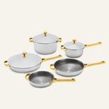 The Titanium Pro Cookware Set+ 2.0 includes a large pot, a saucepan, two frying pans, and a lidded skillet, all featuring silver-toned metal bodies with gold-toned handles and lids.