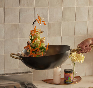 A hand tosses colorful vegetables in a black wok over a stove; some veggies are airborne. A candle, a small vase with yellow flowers, and a spice jar sit on the counter nearby.