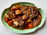 A green oval plate with braised beef short ribs, carrots, and potatoes in a rich brown sauce, garnished with grated horseradish, on a white checkered tablecloth.