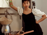 A person in a black dress with white puffed sleeves stands in a kitchen, leaning against a shelf. A pan with food and a wooden spatula sits on the stove, with kitchen utensils and baskets in the background.