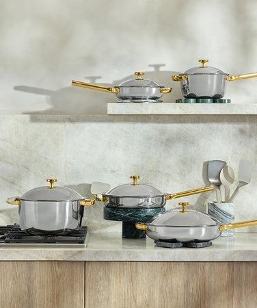 Two silver and gold frying pans with glass lids sit on a stovetop. A brown and white kitchen towel hangs nearby, and utensils are stored in a dark holder on the countertop, with plants in the background.