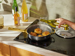 A hand pours white wine from a bottle into a pan of searing chicken thighs on a modern stovetop, with bottles of oil and beverages and a plate with utensils nearby on the counter.
