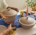 A table set for a meal features two beige pots with lids, a lit candle, a vase with yellow flowers, plates, glasses, and people serving food, creating a warm and inviting atmosphere.